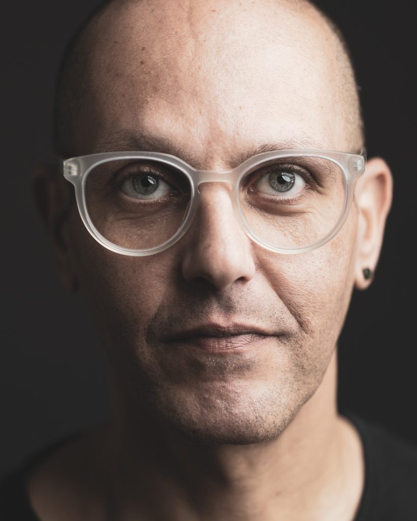 Close-up professional headshot of a man wearing clear glasses against a dark background