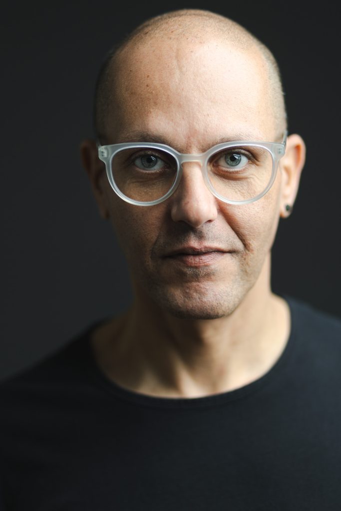 Modern professional headshot of a man wearing clear glasses against a dark background